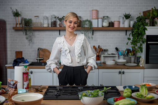 Young Caucasian woman standing and smiling at the kitchen. She is looking at the camera with her hands inside her pocckets. She has vegetables and ingredients for preparing a salad.