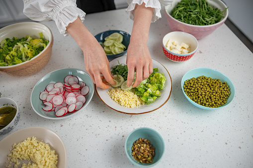Aerial view of the hands of a young Caucasian woman adding cucumber slices to a salad plate with chopped lettuce, arugula and couscous. Around the plate there are many bowls with fresh ingredients like: raddish slices, chopped lettuce, cucumber slices, couscous, green peas, arugula, dressing and pistachios.