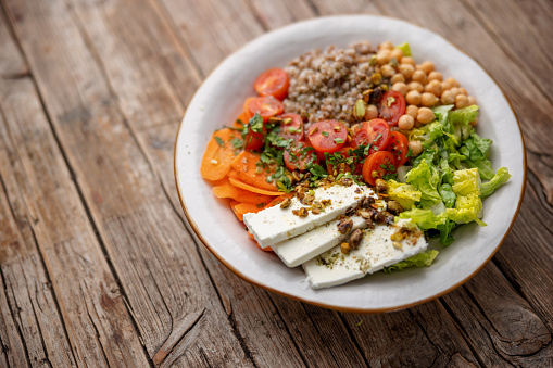 Close-up of a colorful healthy salad containing mixed vegetables, white cheese and buckwheat garnished with chopped herbs and pistachio on the top. Salad is served on a white ceramic plate over a wooden table surface.