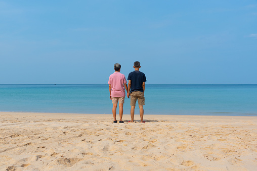 A couple of senior asian adults holding hands and standing on the beautiful beach and blue sky background.