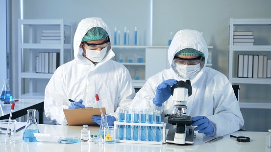 Asian doctor scientist in biohazard protection clothing analyzing virus sample with microscope in laboratory, vaccine research. quarantine or virus outbreak concept