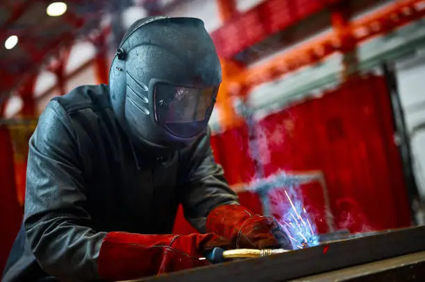 Employee in protective mask and gloves welds manually metal carcass parts in shielding gases at production plant close view