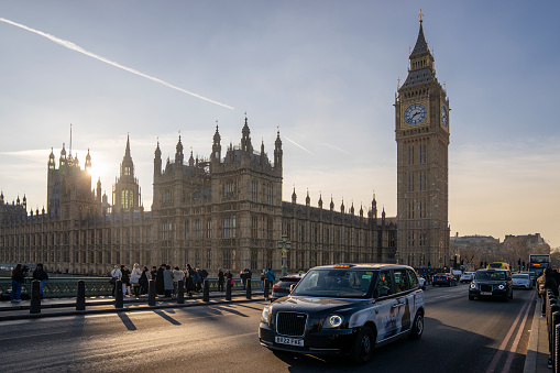 London, United Kingdom - January 22, 2023: Taxi passes Big Ben clock tower and the Houses of Parliament in Westminster, with the sun low in the sky.