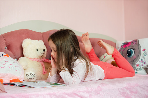 a little girl of five years with long hair lies on her children's cozy bed with toys and reads a book, looks at bright pictures