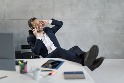 Cheerful mid adult businessman talking over phone with feet up in the office.