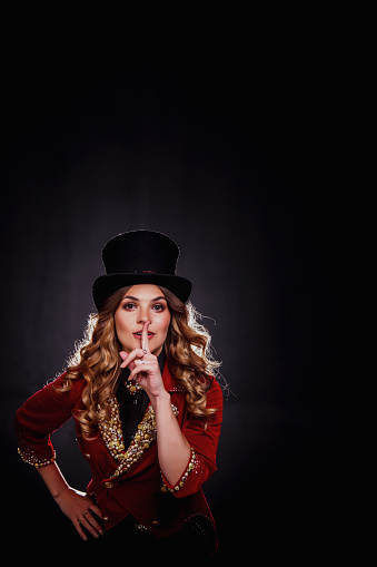 Magician woman with index finger at lips, an illusionist in circus clothes at black background. Actress lady in stage costume showing performance. Concept of fun perform. Copy ad text space, banner