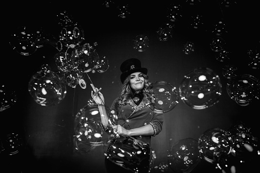 Magician lady make soap bubbles show, an illusionist in stage clothes at black background, black white image. Woman actress in theatre costume. Concept of stage perform and circus. Copy ad text space