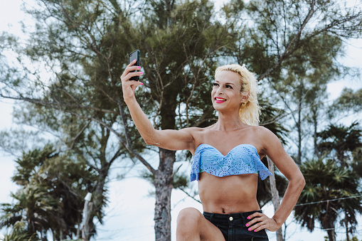 Young transgender woman wearing makeup and woman clothes taking photo selfie with mobile phone at the beach in Mexico Latin America