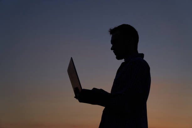 istock Male programmer of IT guy in silhouette using laptop outdoor at sunset, working 1471380010