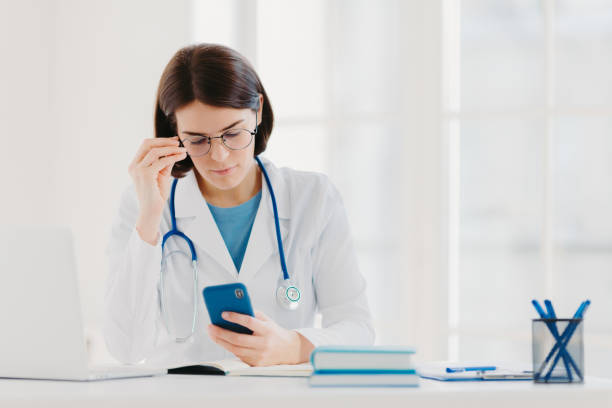 concept de technologie médicale. le médecin de femme concentré regarde l'appareil de smartphone, vérifie l'information nécessaire dans internet, porte le stéthoscope autour du cou, de grandes lunettes rondes, pose dans la clinique. - service computer training office photos et images de collection