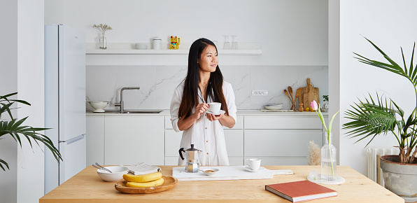 Portrait of a beautiful and happy asian girl in the modern clean minimal white kitchen, early bird morning routine, drinking coffee and looking through the window, representing positive attitude and a healthy lifestyle, an wide image with a large copy space