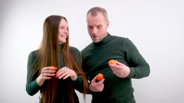 young girl put Tangerines to eyes and pulled out tongue stuck out and she twists and makes faces in the background a man on a white background a green sweater long hair joke fooling around grimacing