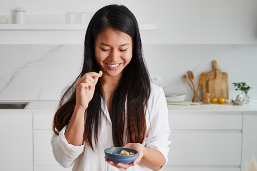 Portrait of a beautiful and happy asian girl in the kitchen, early bird morning routine, eating healthy nuts, representing positive attitude and a healthy lifestyle, an image with a large copy space