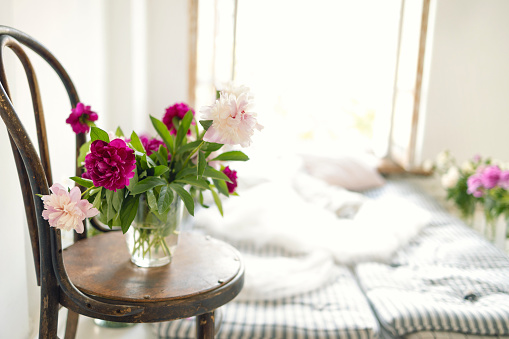 Close up of peonies in a vase on a vintage wooden chair. Rustic style interior with mattresses, pillows and flowers near an open window. Atmospheric moments. Close up.