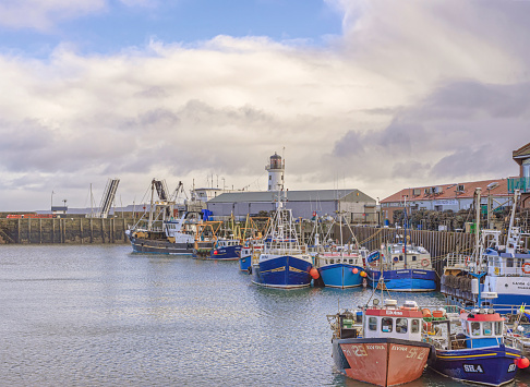 Scarborough, UK. February 24, 2023.  A harbour with trawlers moored to a wharf. A lighthouse is visible over a rooftop and a cloudy sky is above.