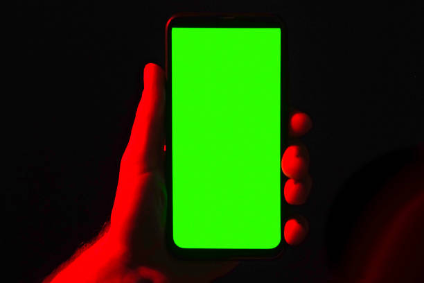 istock Mobile phone in young man hand. Use smartphone with green screen on red blue neon lighting, dark background. Male showing black phone chroma key display. Close up. Hand lifts device up holds it still 1471395415