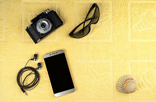 A smartphone with a blank screen, sunglasses, vintage film camera, earphones, and a shell in the background of a yellow beach terry towel.