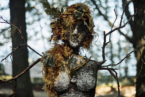 Creepy witch statue art object on forest background made of mud plaster and dry grass, mystical mysterious gloomy statue of woman child of nature. Female scary recycling sculpture in woods