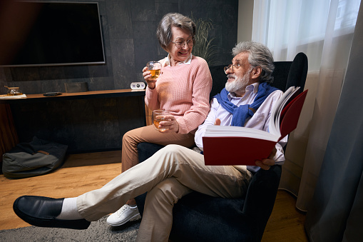 Old spouses are comfortably seated in an armchair with a book and drinks, people communicate pleasantly