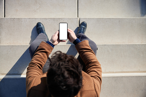 Top view of man in coat sitting on stairs outside holding smartphone with blank white screen