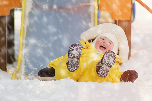 Toddler baby boy rides down a slide playing on a winter playground. A child in a yellow jumpsuit on a children's slide in the snow. Kid age one year eight months