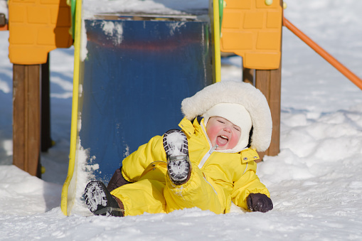 Toddler baby boy rides down a slide playing on a winter playground. A child in a yellow jumpsuit on a children's slide in the snow. Kid age one year eight months