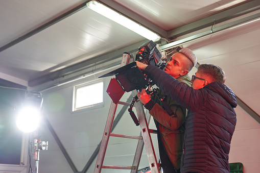 film set with a powerful monoblock lighting fixture and two caucasian men on a ladder ladder under the ceiling with a professional movie camera