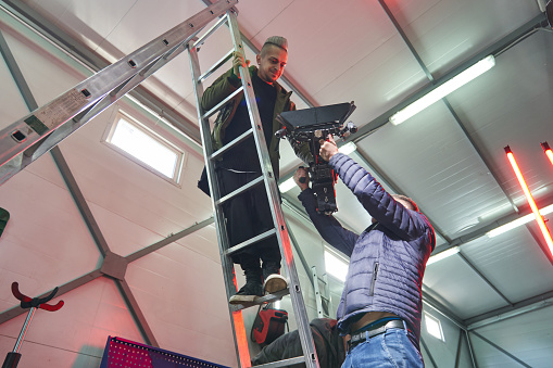 two caucasian men lift a professional movie camera on a high ladder under the ceiling of the film set