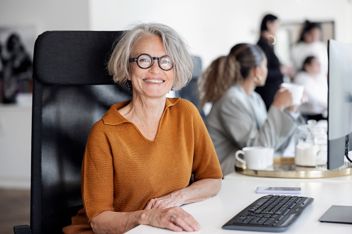 Portrait of a cheerful senior businesswoman sitting at desk with team working in background. Female entrepreneur working at coworking office looking at camera.