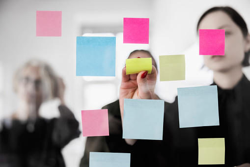 Three businesswoman meeting at office and use post it notes to share ideas. Group of female professionals behind glass using adhesive notes for brainstorming new ideas in office.
