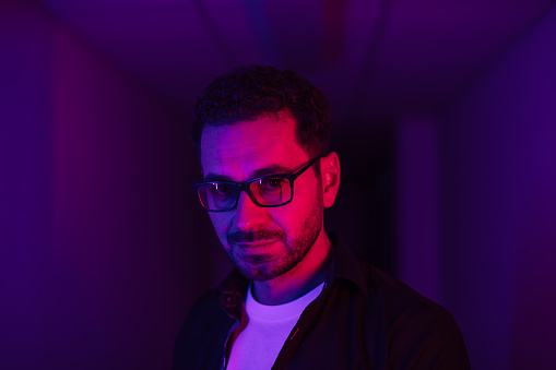 Male portrait of man with neon lighting. Red, blue, purple and magenta colours with dark background. Person wear eyeglasses, dark jacket and white t-shirt indoors. Futuristic lifestyle concept multicolour light