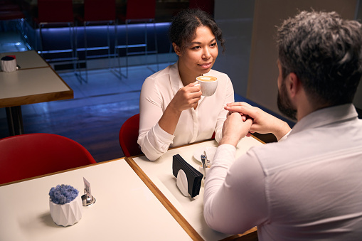 Romantic woman with cup of cappuccino and male holding hands at table in coffee shop