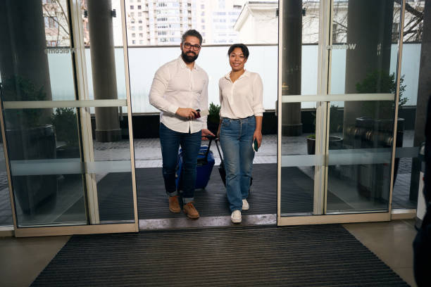 Joyous tourist and his pleased female companion arriving at hotel Full-size portrait of happy couple with travel documents and luggage walking through open sliding doors walking into glass door stock pictures, royalty-free photos & images