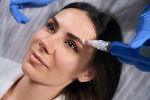 Medical worker in protective gloves holding device and doing post acne treatment to beautiful female in salon