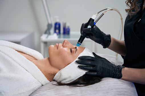 Cosmetologist in protective gloves sitting near beautiful lady and doing Hydradermabrasian procedure in salon