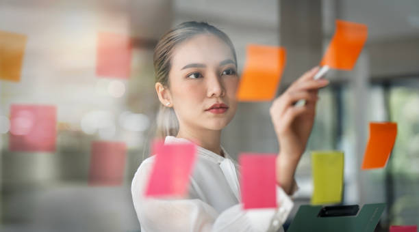 istock Young smiley attractive businesswoman using sticky notes in glass wall to writing strategy business plan to development grow to success 1471431401