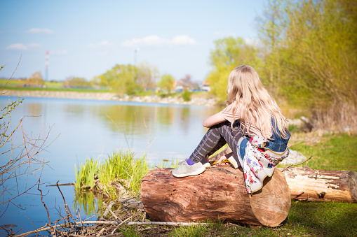 girl with long blond hair sitting by the river in scenic spring nature, back view