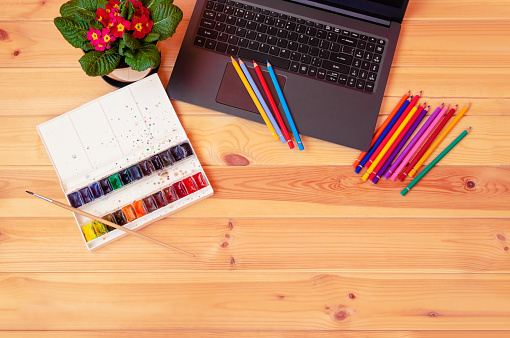 Color pencils, watercolor paints, primrose flower and laptop on desk. Top view, copy space.
