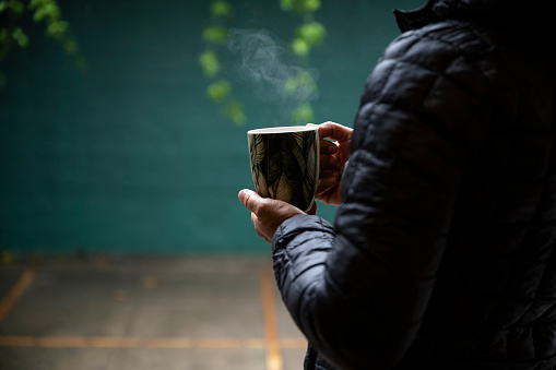 Woman wearing recycled plastic clothing standing by open door looking out at rain in courtyard holding mug of steaming tea