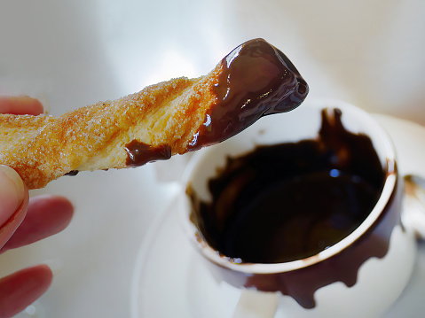 A hand holding a churros stick dipped with hot thick chocolate, ready to eat, Spanish coffee shop, blurred white cup background
