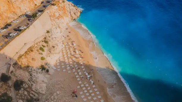 Drone high-angle photo of crystal blue sea with yellow sand at the beautiful beach hidden in the cliff with cars driving the mountain pass in Turkey, Middle East