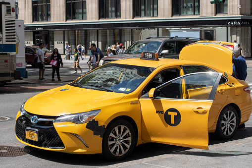 New York, NY, USA - July 4, 2022: A taxi driver pulls his yellow cab over on the streets in Midtown Manhattan, New York City, and helps the passenger get the luggage from the trunk.