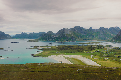 A couple of travelers getting to the top of the mountain, admiring the view of the turquoise-coloured sea, green meadow, and dramatic peaks in Northern Norway