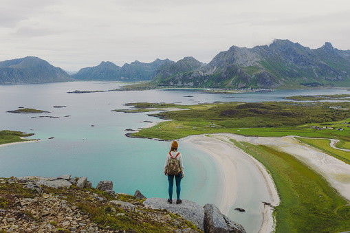Rear view of woman traveler in a yellow hat getting to the top of the mountain, admiring the view of the turquoise-coloured sea, green meadow and dramatic peaks in Northern Norway