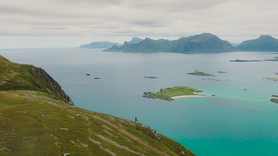 A couple of travelers getting to the top of the mountain, admiring the view of the turquoise-coloured sea, green meadow, and dramatic peaks in Northern Norway