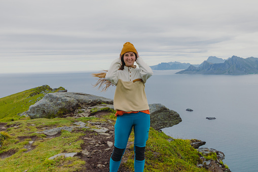 Front view of smiling woman traveler in a yellow hat feeling freedom on the top of the mountain, admiring the view of the turquoise-colored sea, green meadow, and dramatic peaks in Northern Norway