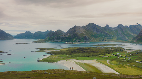 A couple of travelers getting to the top of the mountain, admiring the view of the turquoise-coloured sea, green meadow, and dramatic peaks in Northern Norway