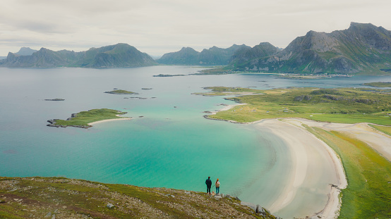 A couple of travelers getting to the top of the mountain, admiring the view of the turquoise-coloured sea, green meadow, and dramatic peaks in Northern Norway