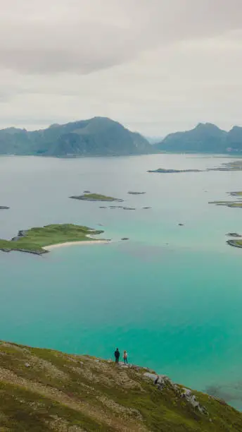 Photo of Aerial view of woman and man contemplating scenic view of the ocean and mountains from top of Lofoten Islands