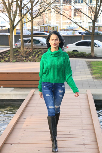 An Indian model practicing her runway walk outdoors. She is wearing long black straight hair, a green sweater, torn jeans and black knee high boots.